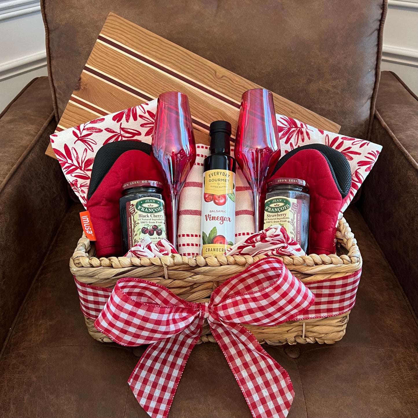 Red-themed gift basket with vibrant kitchen linens, gourmet jams, balsamic vinegar, and a handcrafted wood board made of cherry, maple, and padauk.