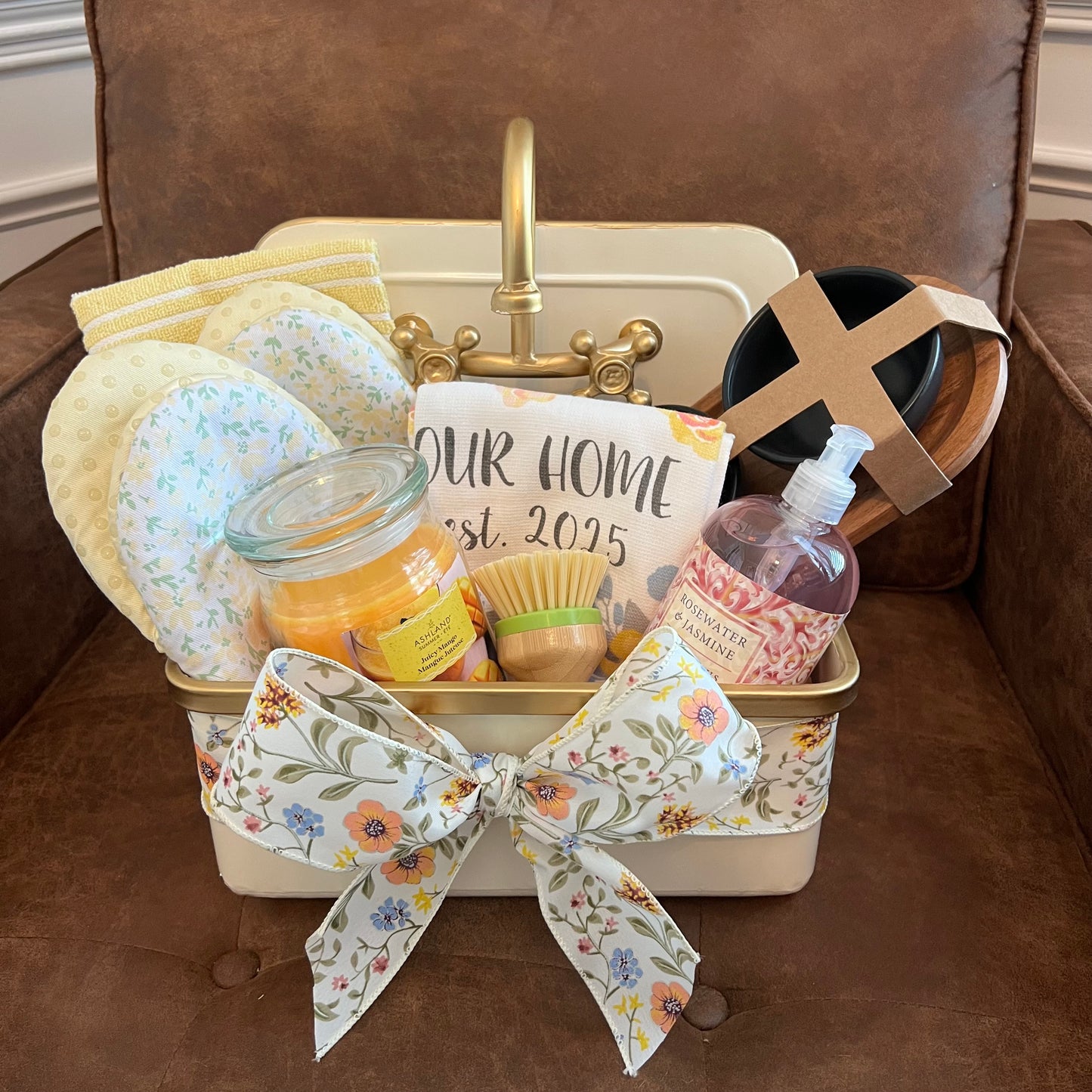 Housewarming gift basket styled inside a decorative sink, filled with oven mitts, hand soap, candle, towels, scrubber, and a three-bowl flight board.