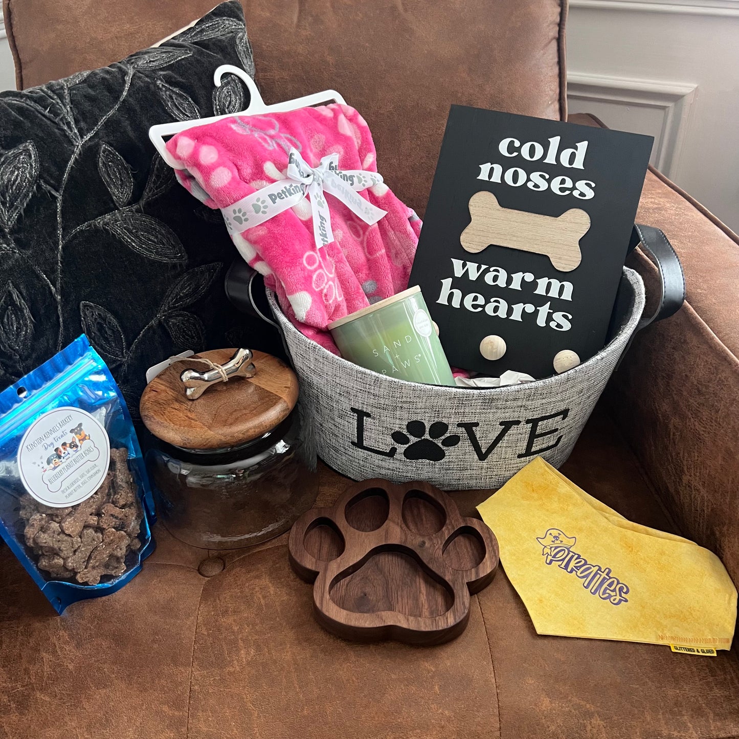 Dog-themed gift basket on a couch including a pink blanket, “Cold Noses Warm Hearts” leash holder, glass treat jar with wood lid, Sand + Paws candle, handmade yellow Pirates dog bandana, wooden paw-shaped dish, and a bag of blueberry peanut butter dog treats from Kinston Kennels. (2)