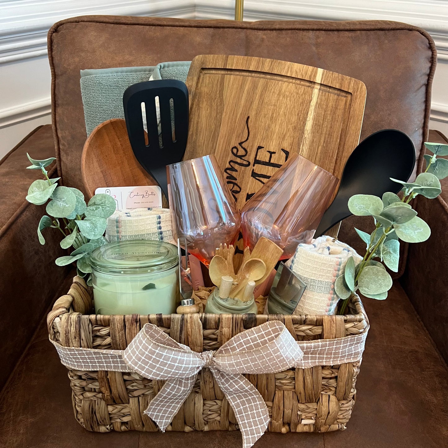 Welcome Home gift basket with engraved charcuterie board, candle, towels, spoon, wine glasses, cheese tools, and greenery