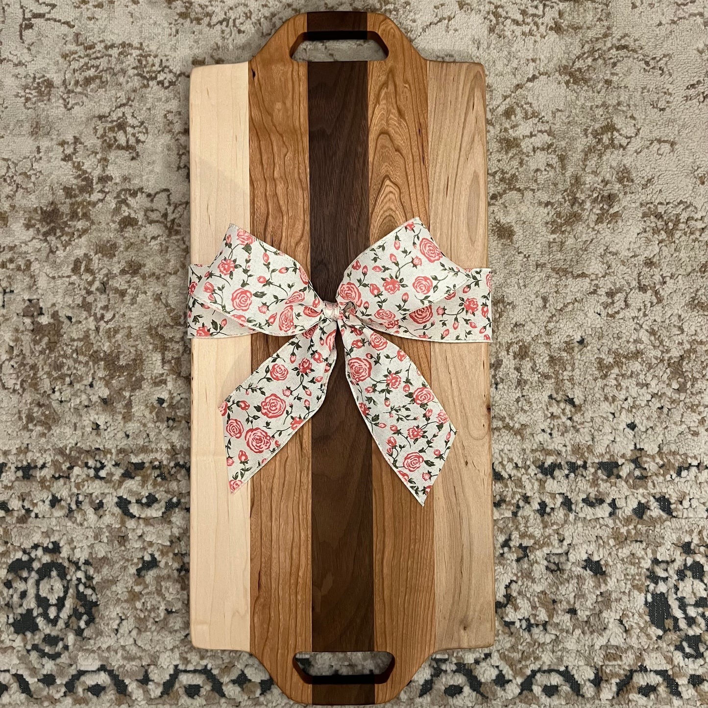 Handcrafted charcuterie board made from walnut, maple, and cherry wood, featuring dual handle cutouts and a floral gift bow, placed on a textured rug background.”