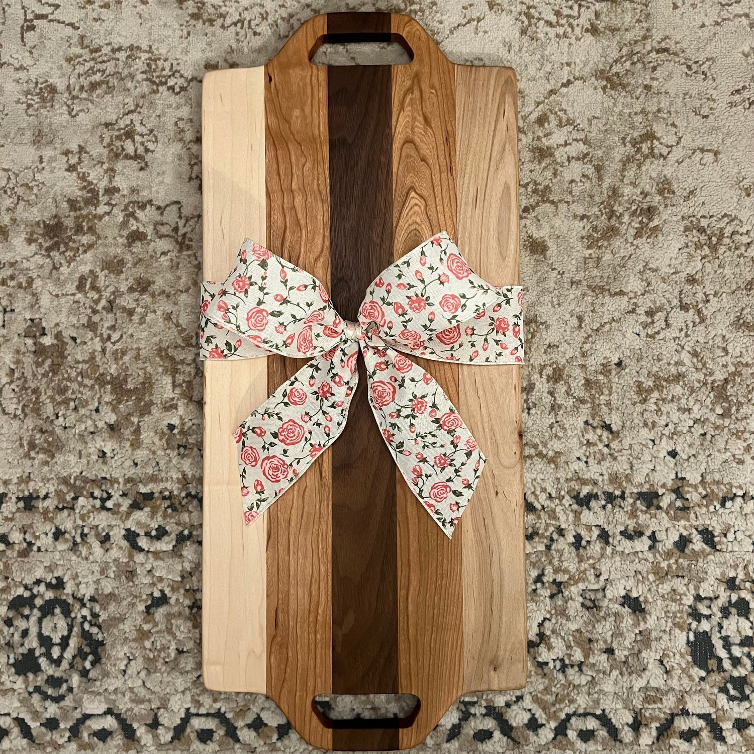 Handcrafted charcuterie board made from walnut, maple, and cherry wood, featuring dual handle cutouts and a floral gift bow, placed on a textured rug background.”