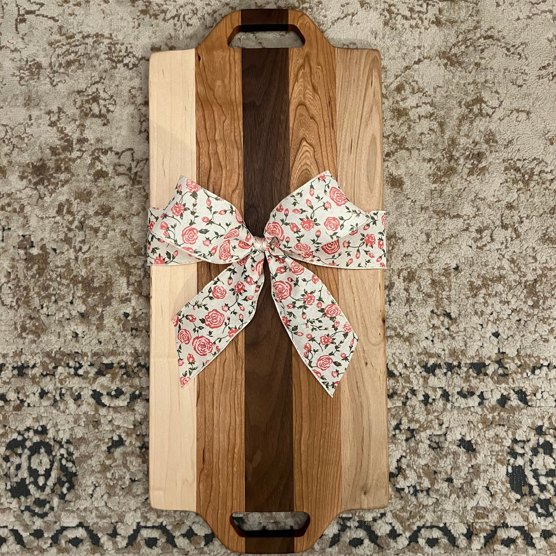 Handcrafted charcuterie board made from walnut, maple, and cherry wood, featuring dual handle cutouts and a floral gift bow, placed on a textured rug background.”