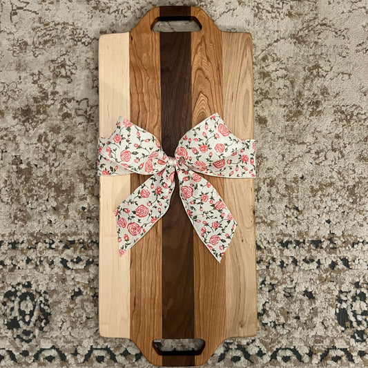 Handcrafted charcuterie board made from walnut, maple, and cherry wood, featuring dual handle cutouts and a floral gift bow, placed on a textured rug background.”