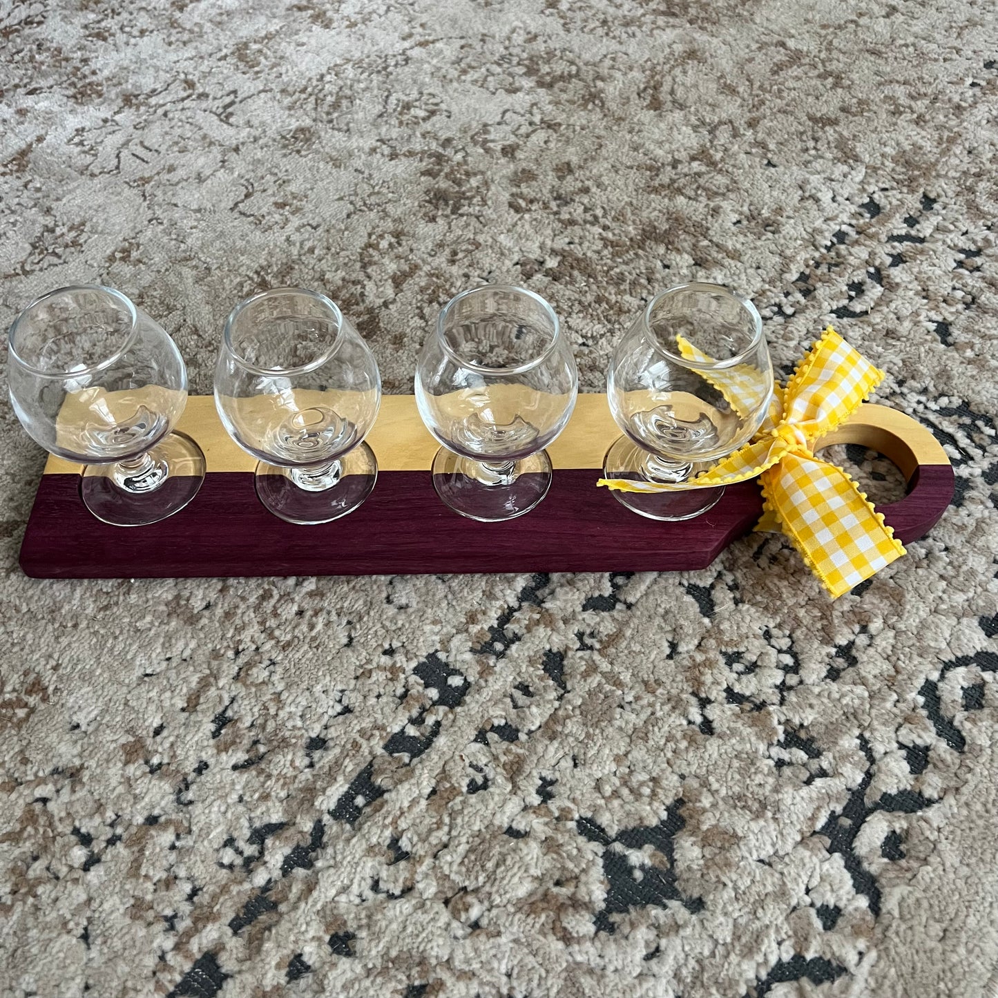 Handcrafted bourbon flight board made from purpleheart and yellowheart wood, featuring four tasting glasses arranged on a vibrant wood grain surface.