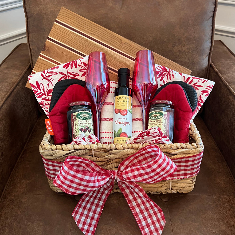 Red-themed gift basket with vibrant kitchen linens, gourmet jams, balsamic vinegar, and a handcrafted wood board made of cherry, maple, and padauk.