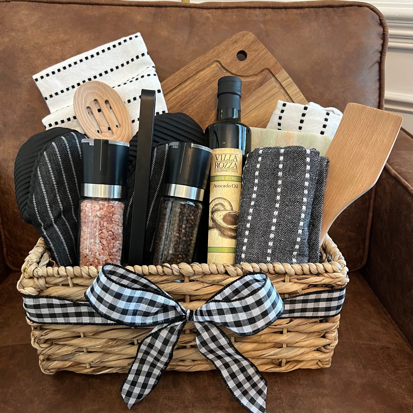 Farmhouse kitchen gift basket with marble cheese board, utensils, towels, and gourmet ingredients in black and neutral tones.