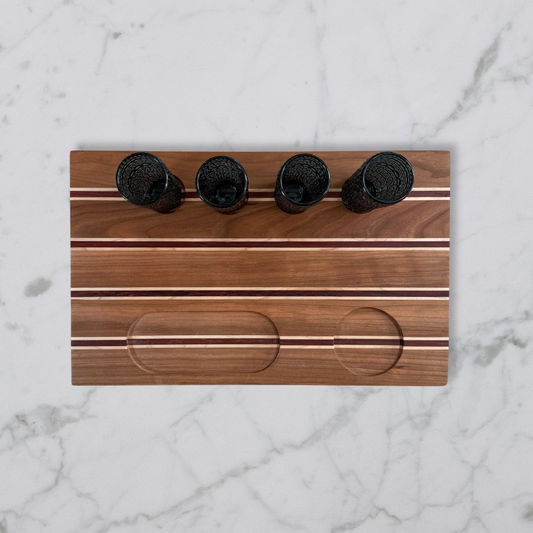 Handcrafted tequila flight board with cherry, maple, and padauk wood and four shot glasses on marble surface