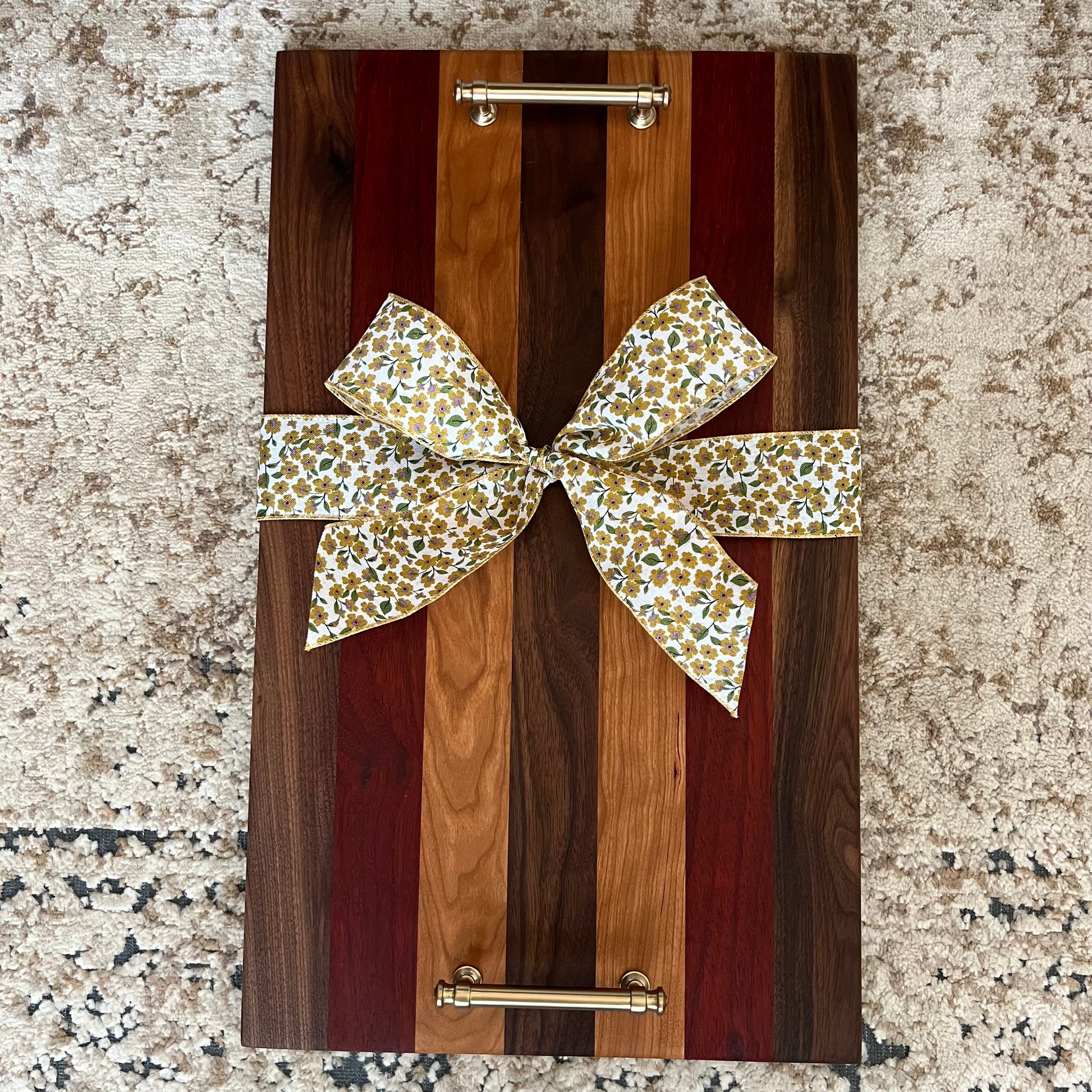 Handcrafted wood tray made from walnut, padauk, and cherry with gold handles and a floral bow, displayed on a textured rug.