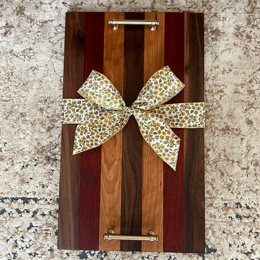 Handcrafted wood tray made from walnut, padauk, and cherry with gold handles and a floral bow, displayed on a textured rug.