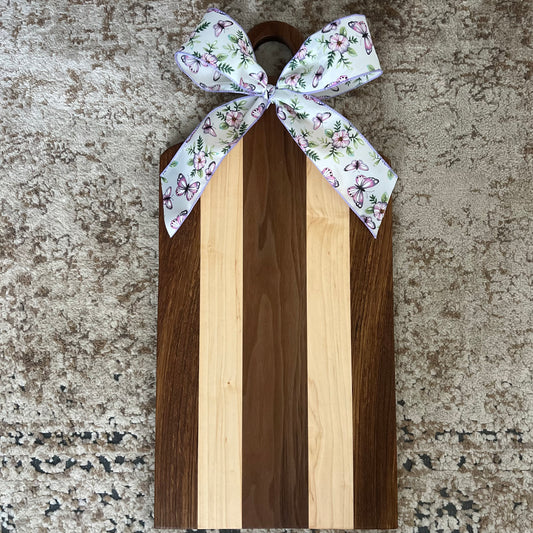 Handled walnut and cherry charcuterie board with a floral bow, displayed on a neutral rug.