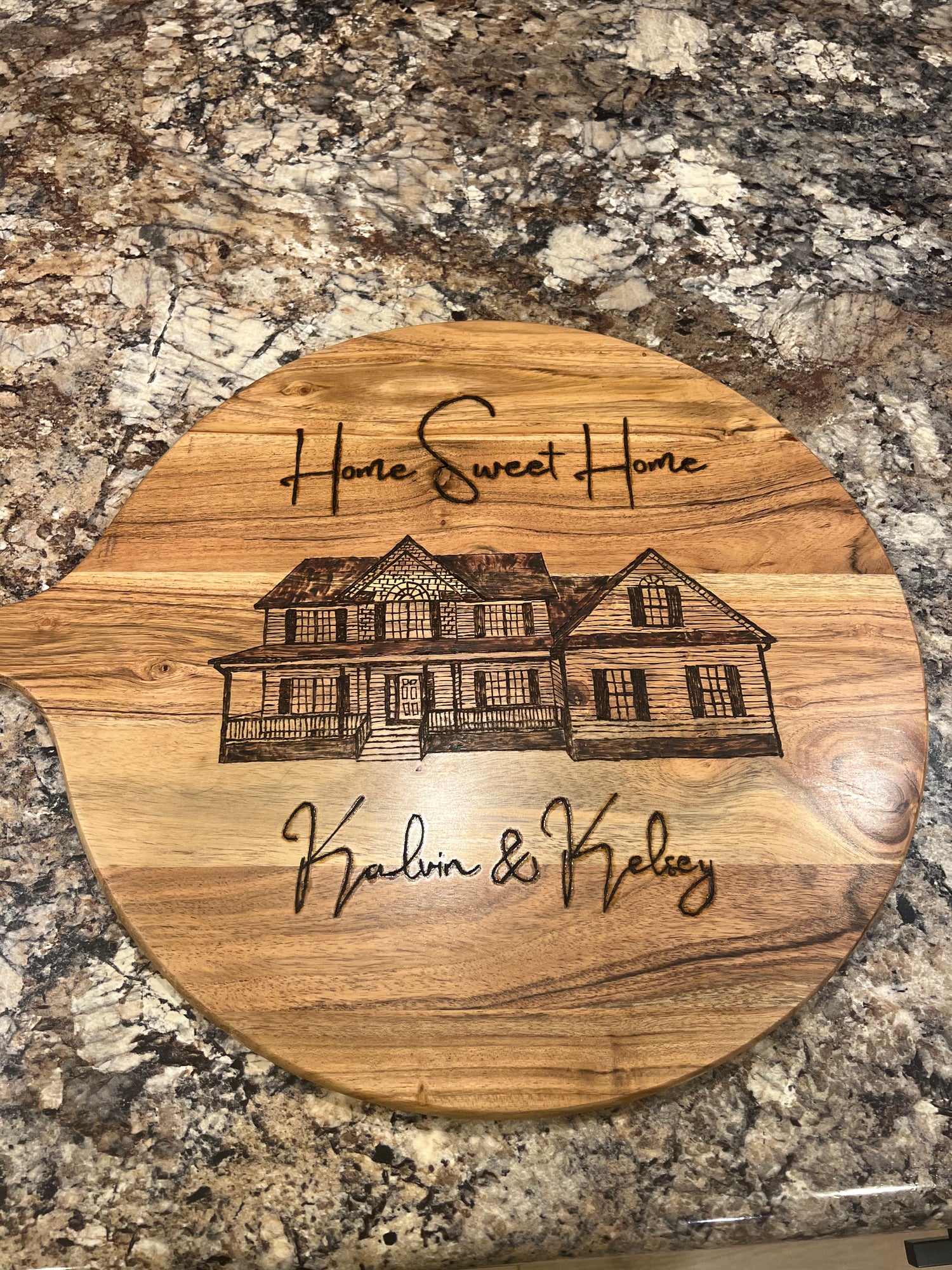 Handcrafted round wooden board with a custom wood-burned illustration of a home, featuring the phrase “Home Sweet Home” and personalized names “Kalin & Kelsey,” accented with a blue patterned bow.