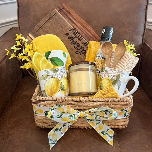 Lemon-themed gift basket with lemon mugs, candle, towels, spoons, sea salt, lemon measuring spoons, and a custom wood-burned "Welcome to Our Home" charcuterie board.