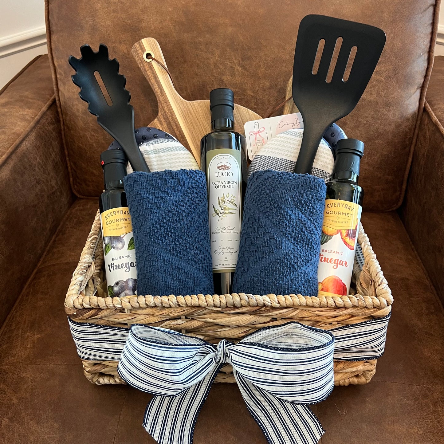 Neutral-toned basket with navy towels, striped bow, olive oil, balsamic vinegars, black utensils, kitchen mitts, and a wood board.