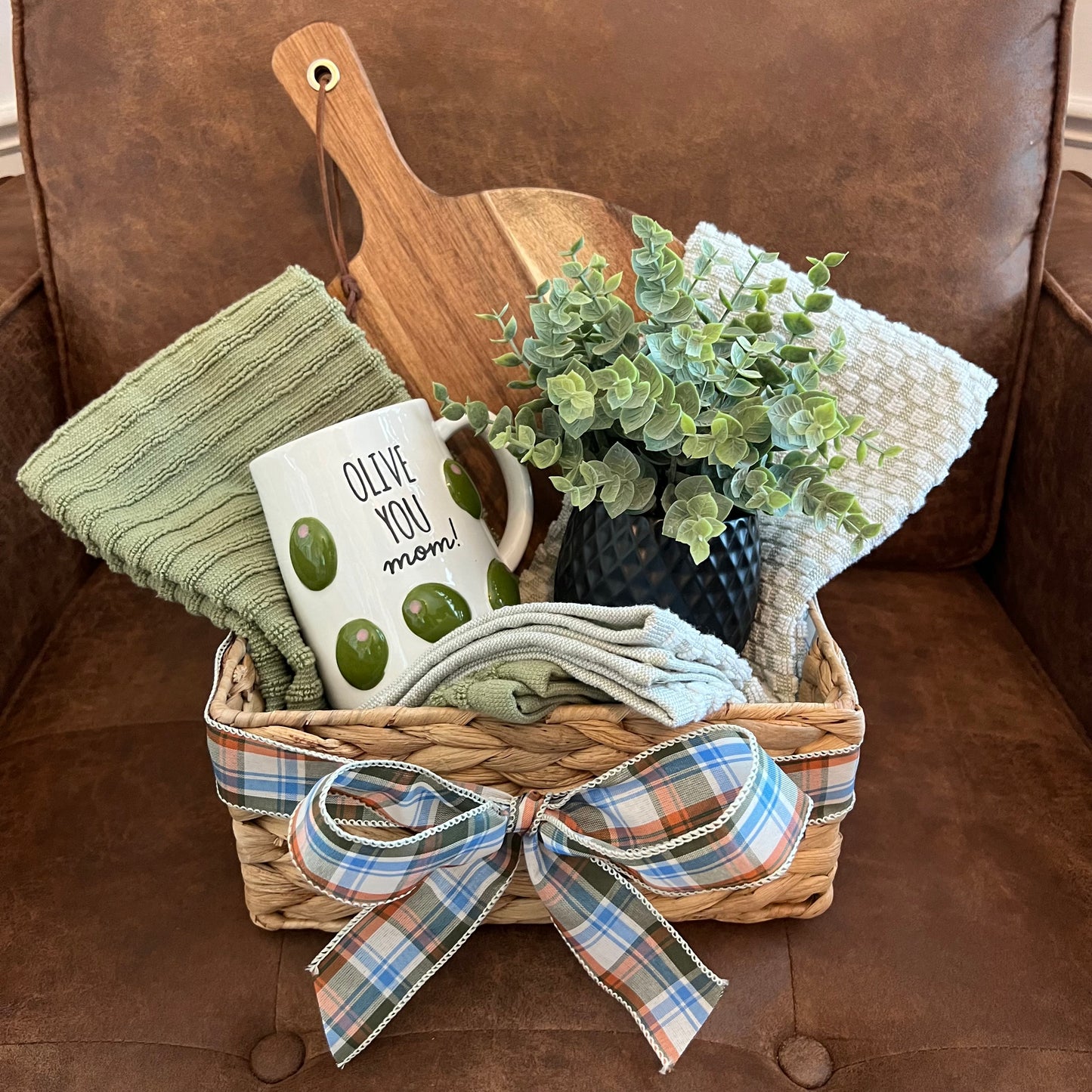 Mother’s Day gift basket with green towels, a mug that reads "Olive You Mom!", a black vase with faux greenery, and a paddle-style wood board.