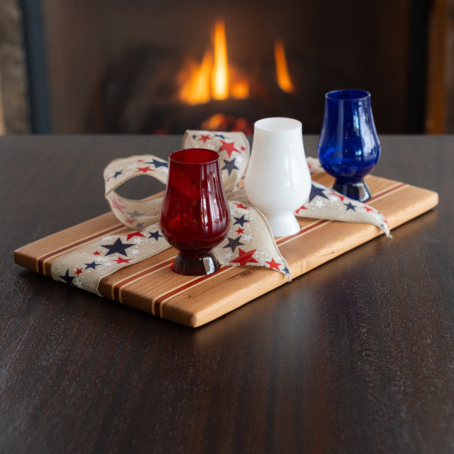 Three colored Glencairn glasses on a patriotic wooden bourbon flight board with a fireplace in the background