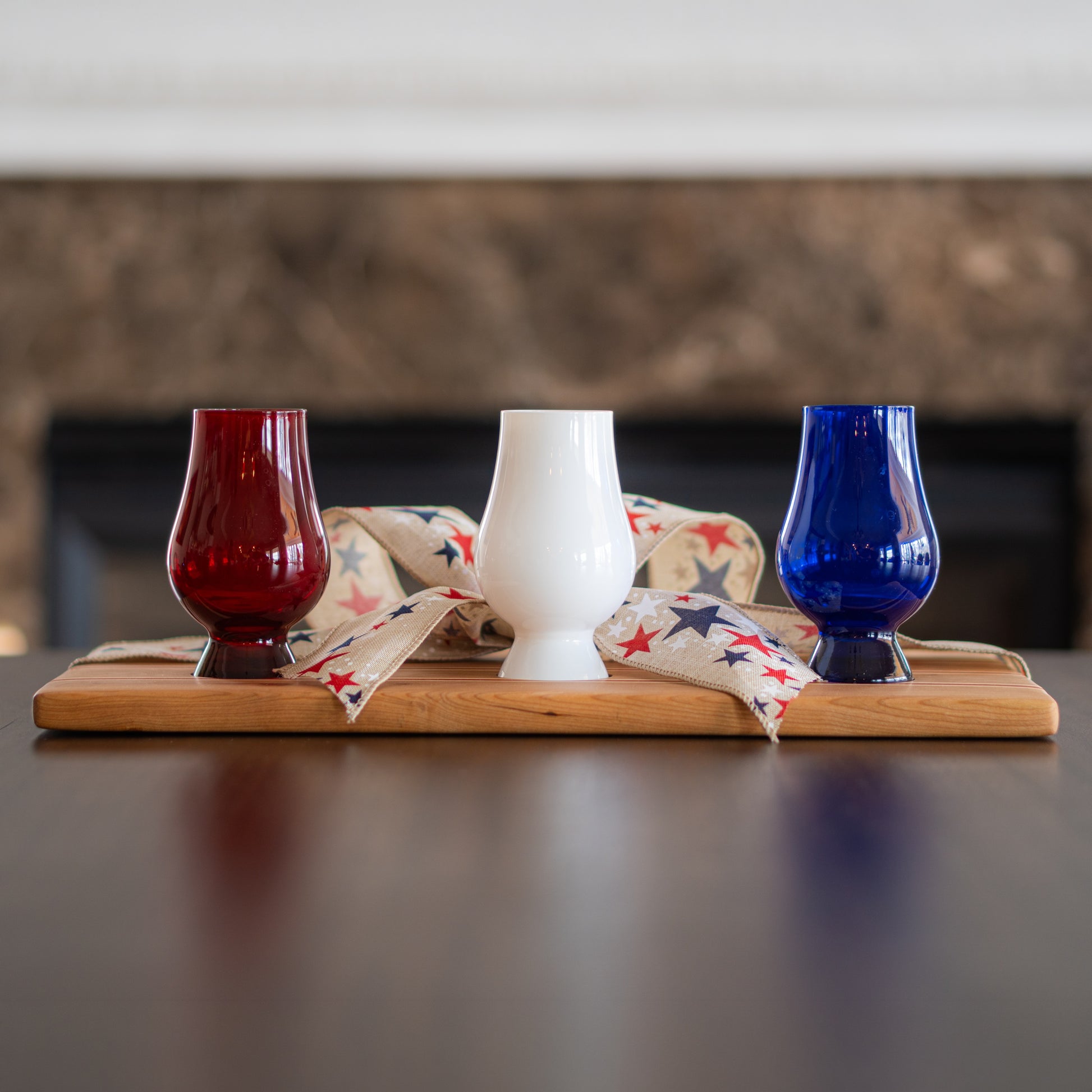 Three colored Glencairn glasses on a patriotic wooden bourbon flight board with a fireplace in the background
