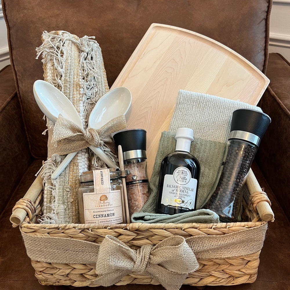 Neutral woven basket filled with a light maple board, ceramic spoons, natural linens, balsamic vinegar, spice grinders, and a cinnamon jar.