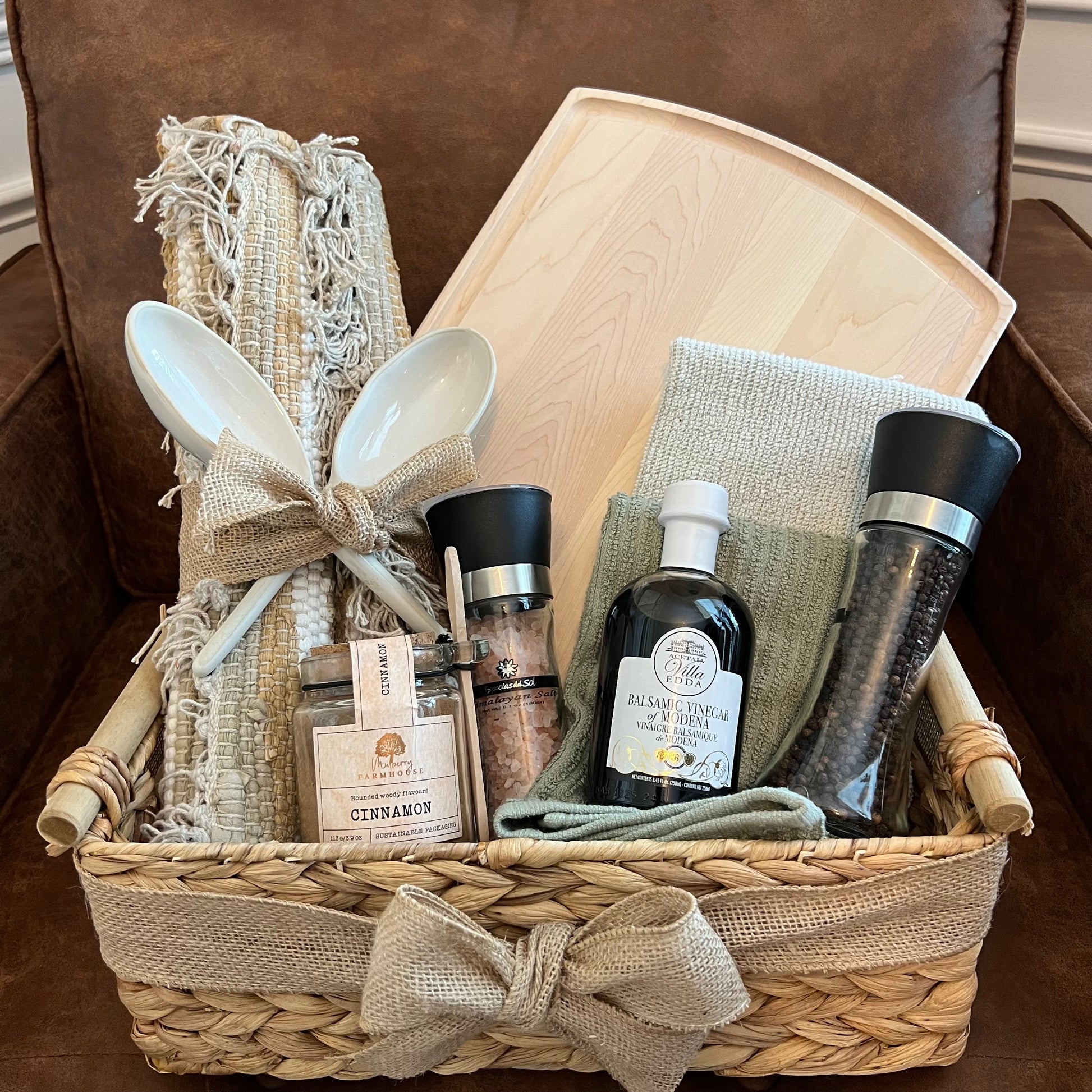 Neutral woven basket filled with a light maple board, ceramic spoons, natural linens, balsamic vinegar, spice grinders, and a cinnamon jar.