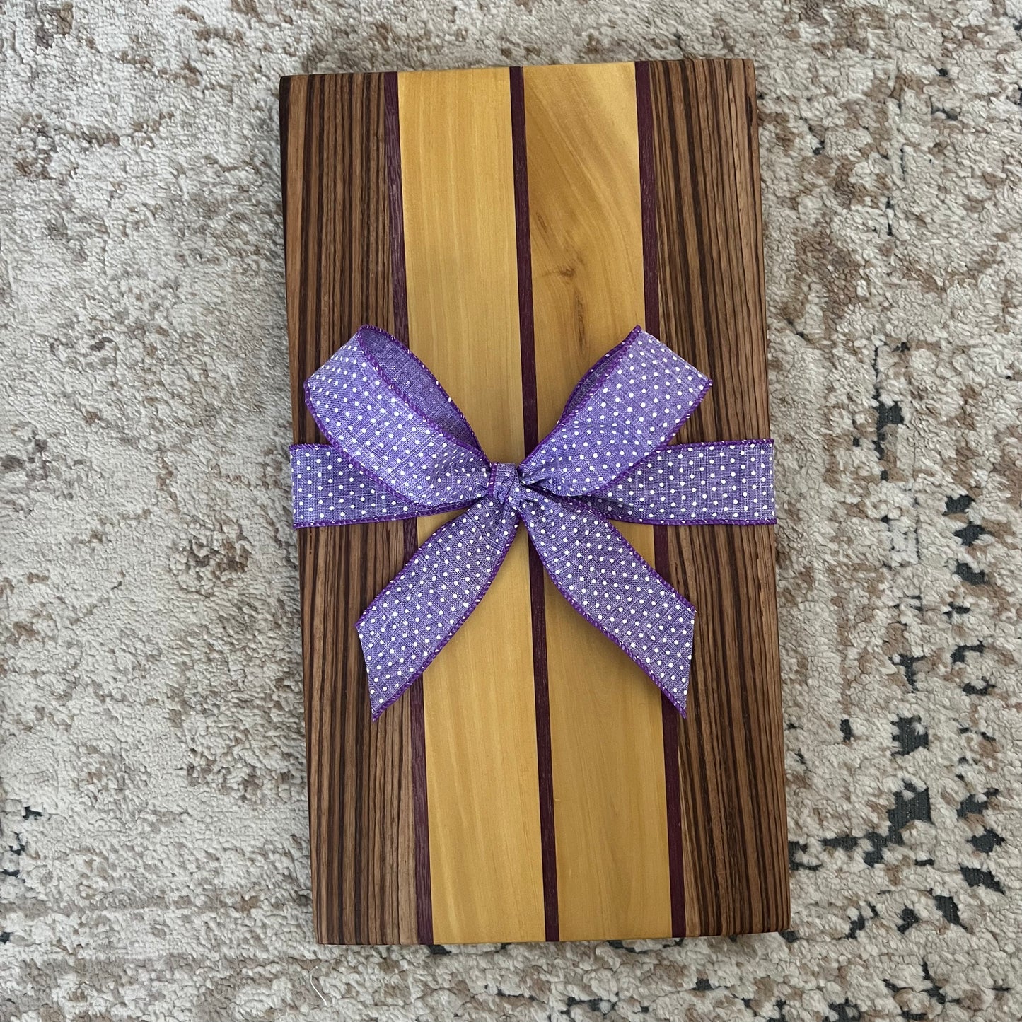 Serving board made from purpleheart, yellowheart, and zebrawood, measuring 16 by 9 inches. Handcrafted with bold natural wood stripes and smooth finish.