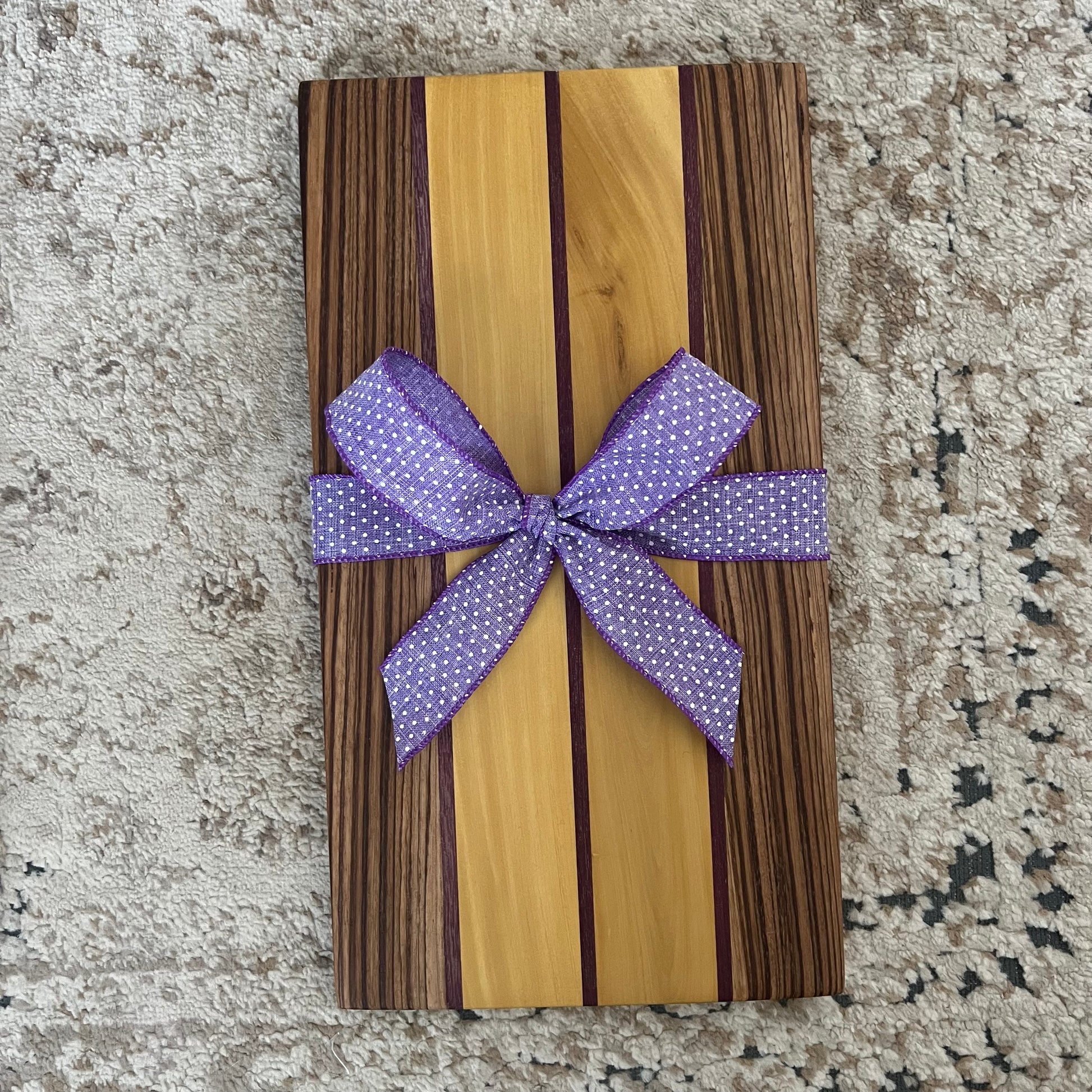 Serving board made from purpleheart, yellowheart, and zebrawood, measuring 16 by 9 inches. Handcrafted with bold natural wood stripes and smooth finish.
