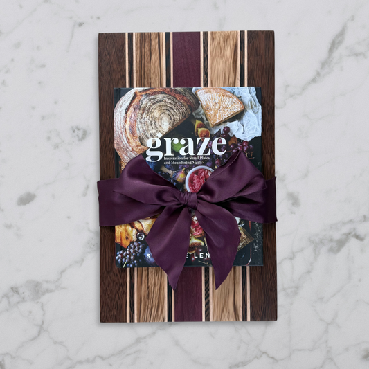 Handcrafted striped wood charcuterie board made from brownheart, purpleheart, maple, zebrawood, and wenge, paired with the Graze book and wrapped with ribbon on a marble surface
