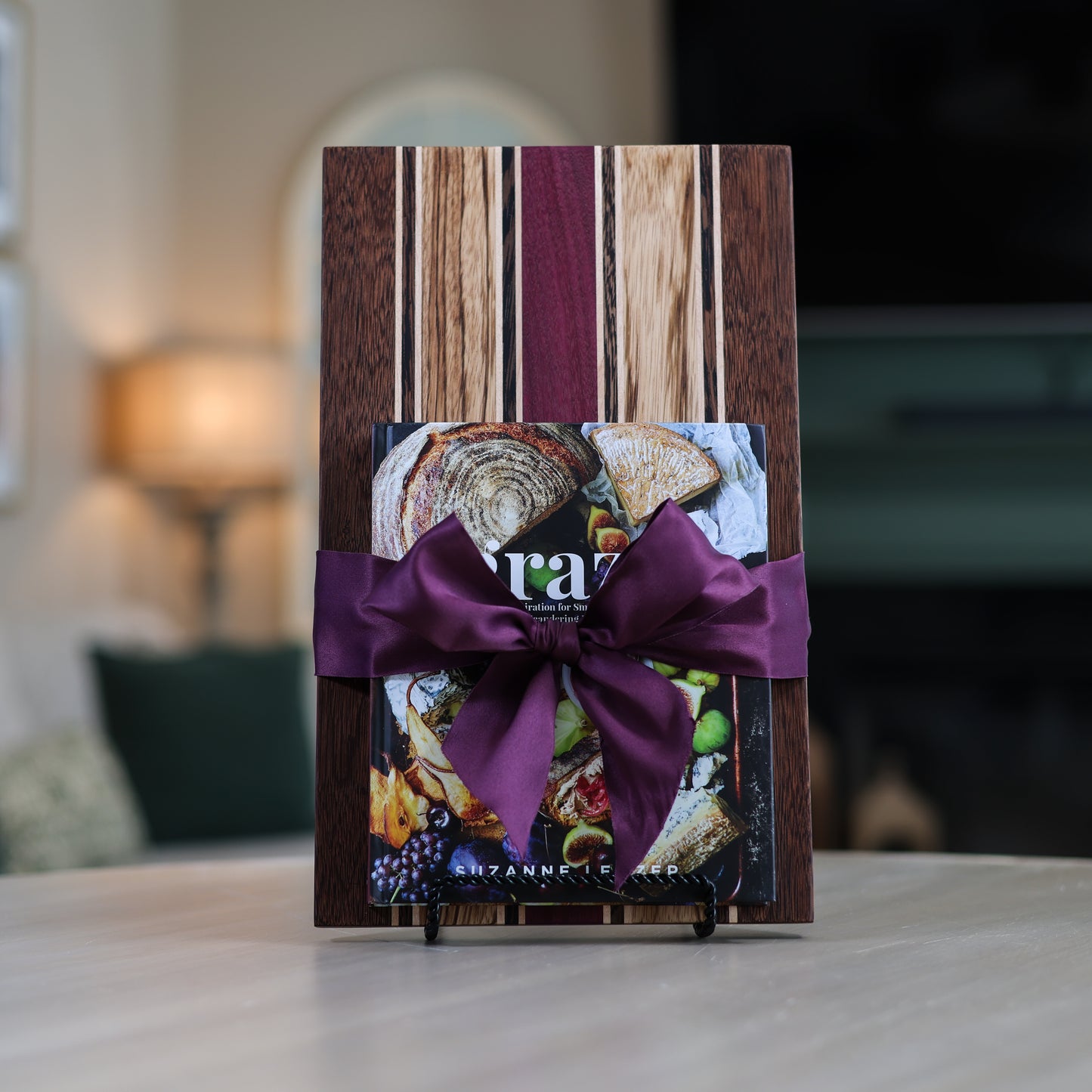 Handcrafted striped wood charcuterie board made from brownheart, purpleheart, maple, zebrawood, and wenge, paired with the Graze book and wrapped with ribbon on a living room table