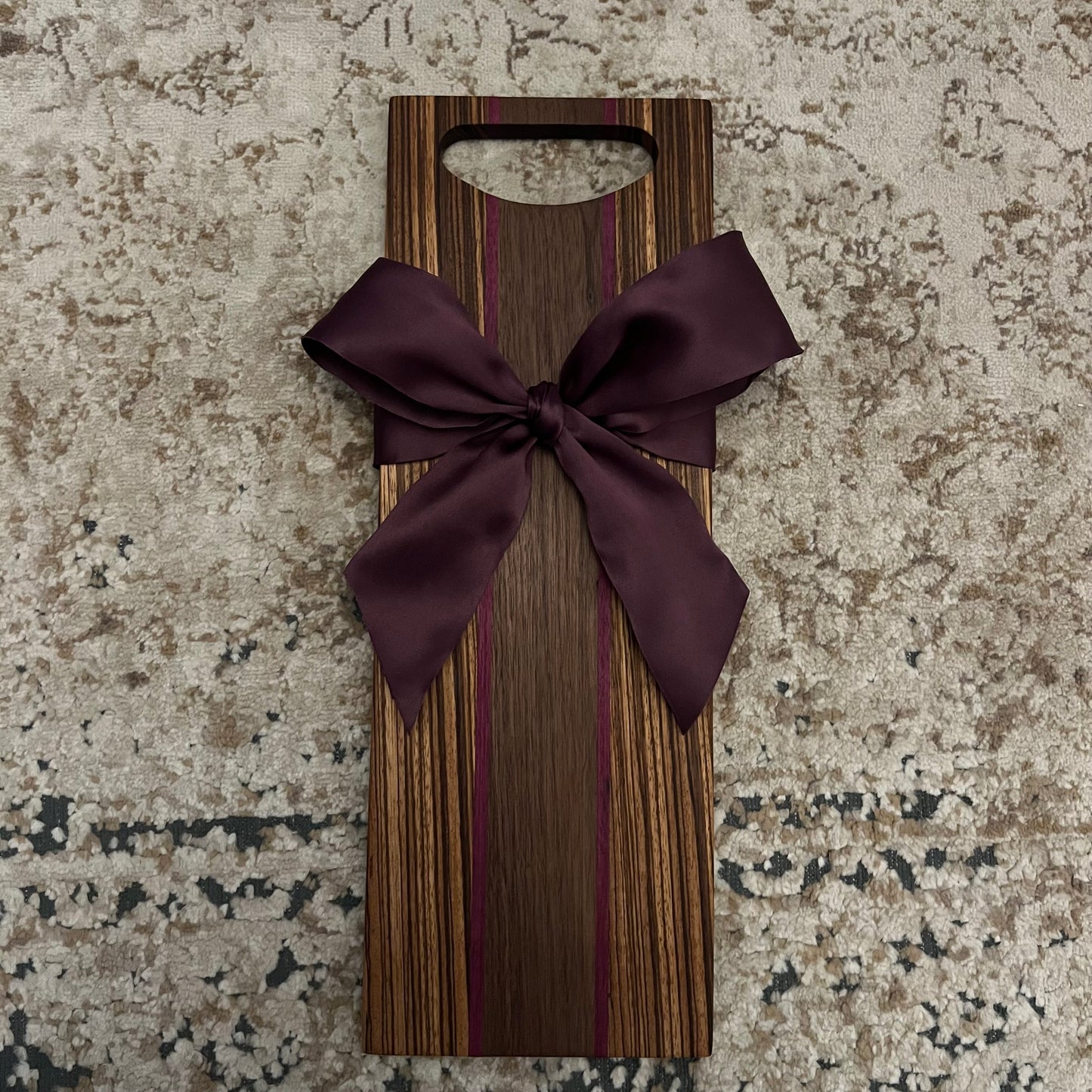Handcrafted zebra wood charcuterie board with walnut and purple heart wood strips, plum bow, and rounded handle, 18.5" x 6.5"