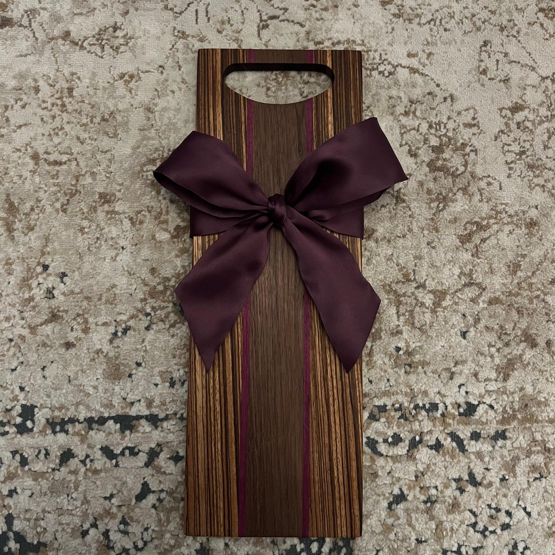 Handcrafted zebra wood charcuterie board with walnut and purple heart wood strips, plum bow, and rounded handle, 18.5" x 6.5"