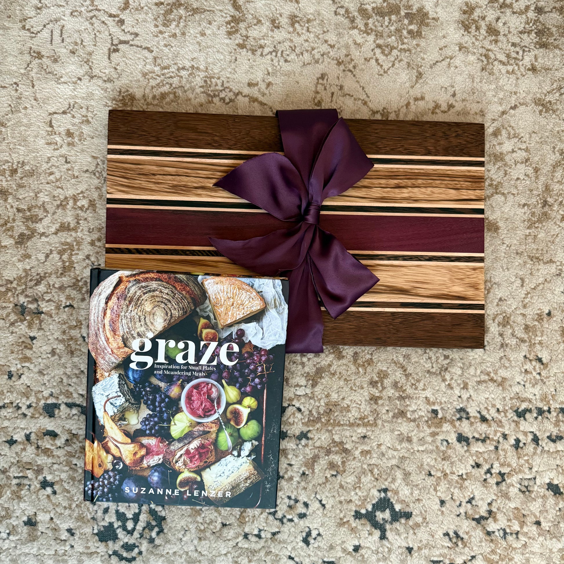 Wooden cutting board with a purple ribbon and a cookbook titled 'graze' on a textured surface.