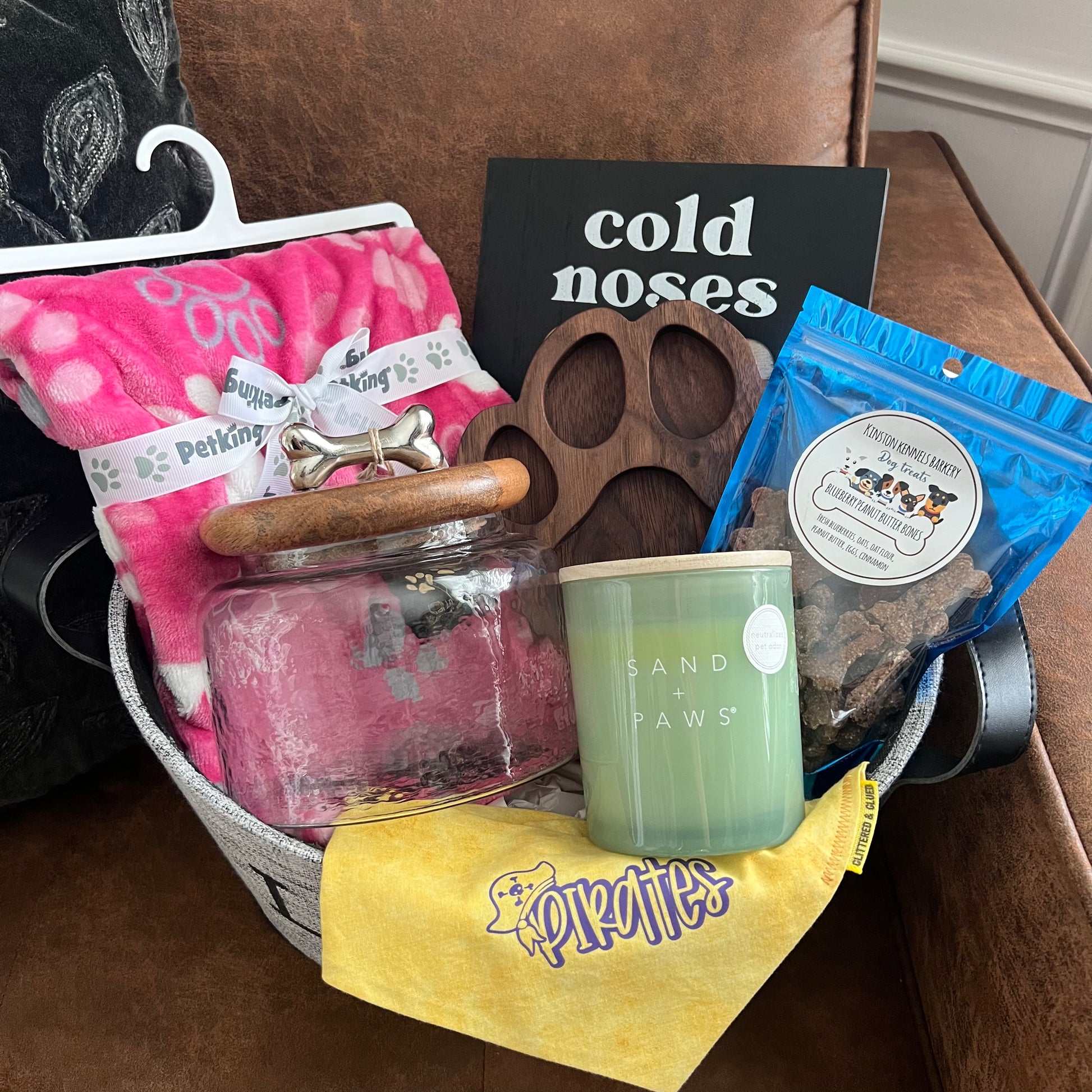 Dog-themed gift basket on a couch including a pink blanket, “Cold Noses Warm Hearts” leash holder, glass treat jar with wood lid, Sand + Paws candle, handmade yellow Pirates dog bandana, wooden paw-shaped dish, and a bag of blueberry peanut butter dog treats from Kinston Kennels.
