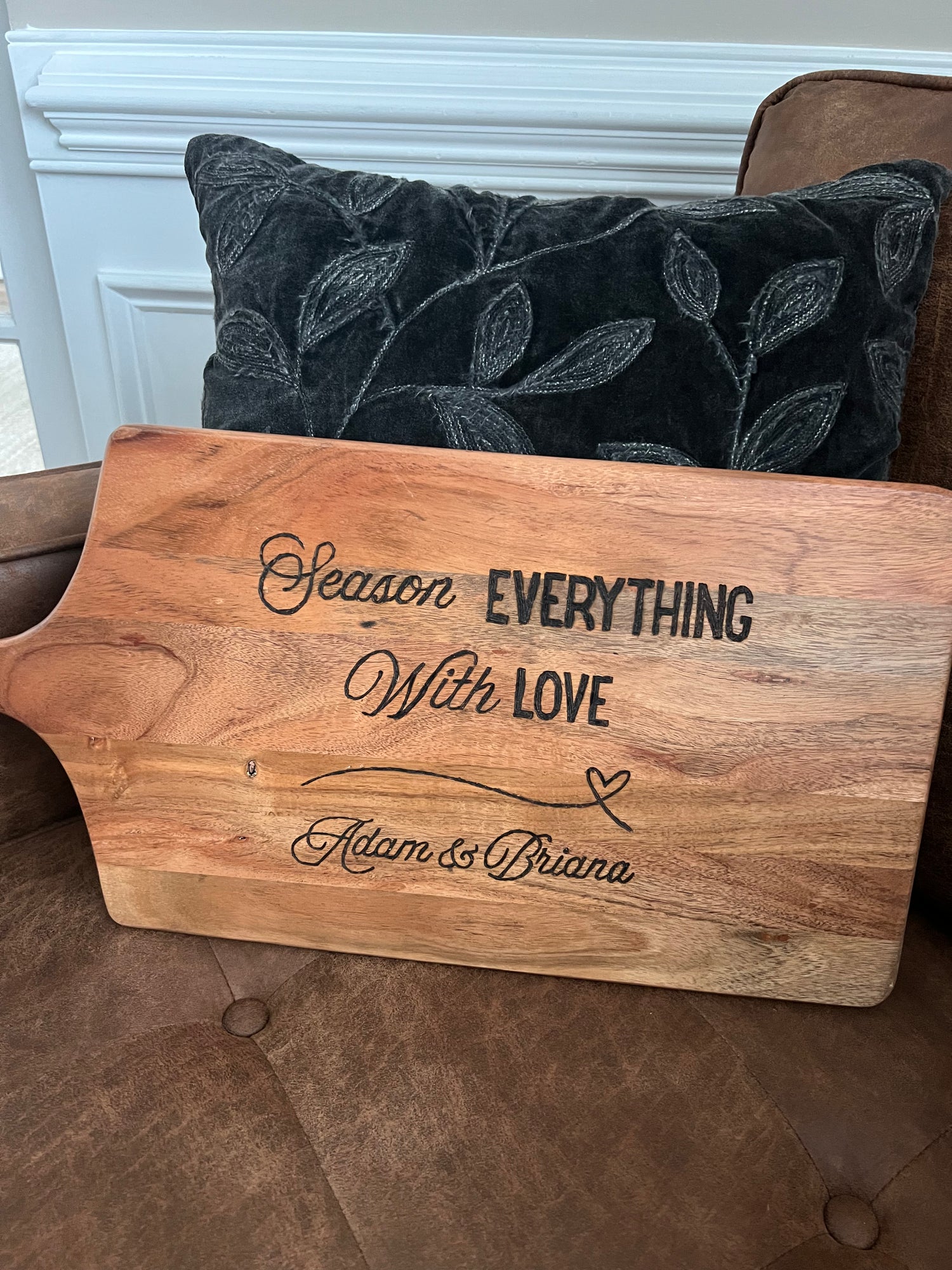 Handcrafted wooden board engraved with the phrase “Season Everything With Love” and personalized with the names “Adam & Briana,” resting on a brown couch with a dark leaf-patterned pillow in the background.