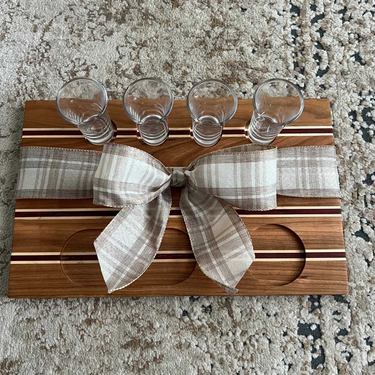 Handcrafted tequila flight board made from cherry, maple, and African padauk wood, featuring four glass shot glasses arranged on a striped wooden board.