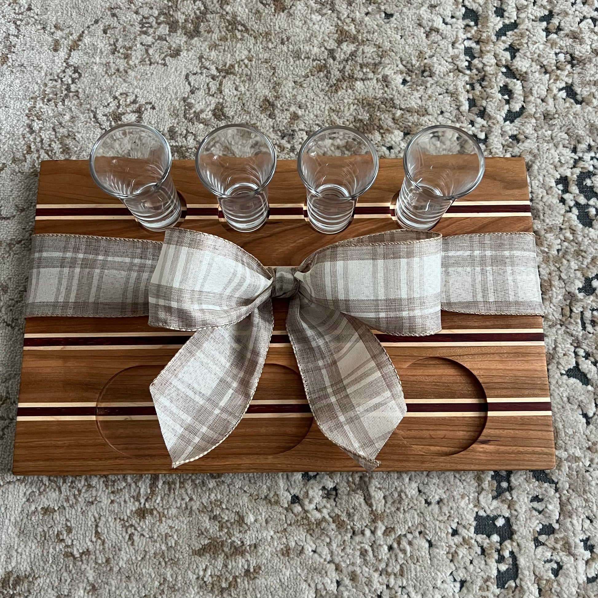 Handcrafted tequila flight board made from cherry, maple, and African padauk wood, featuring four glass shot glasses arranged on a striped wooden board.