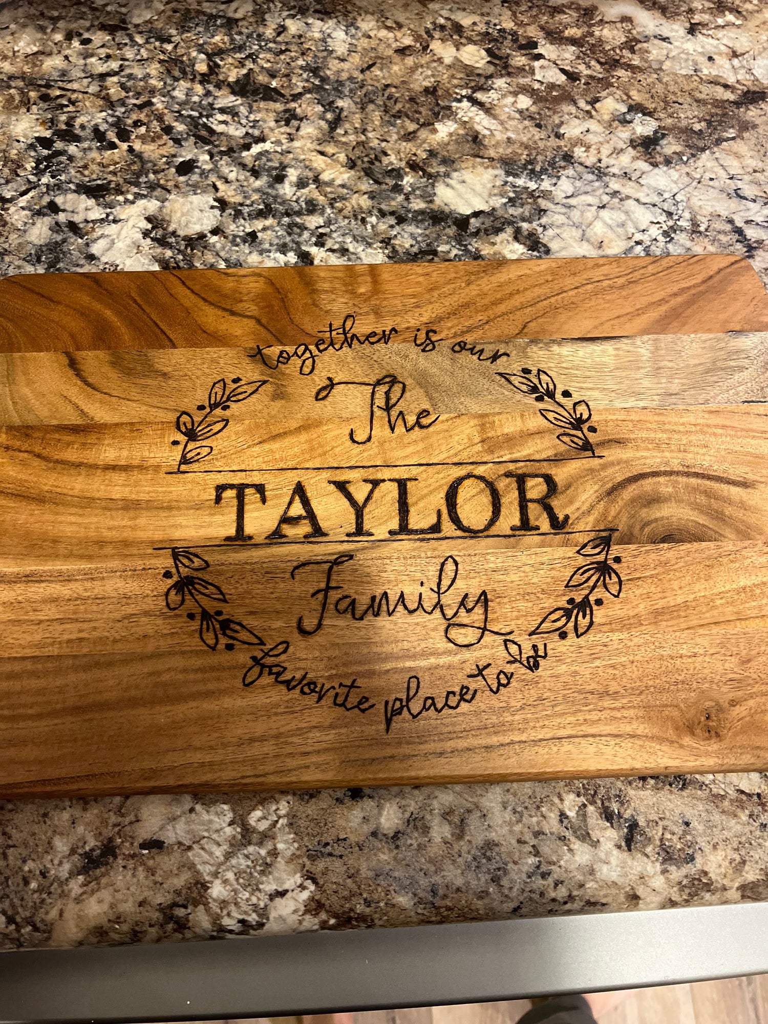 Hand-burned wooden board with the phrase “Together is our favorite place to be – The Taylor Family,” surrounded by leaf accents, displayed on a stone-patterned countertop.