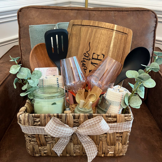 Welcome Home gift basket with engraved charcuterie board, candle, towels, spoon, wine glasses, cheese tools, and greenery
